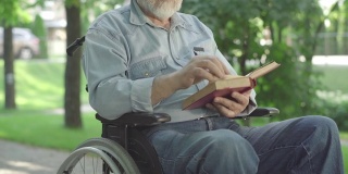 无法辨认的瘫痪老人翻动书页，大声朗读。白发苍苍的退休白人在阳光明媚的夏日公园里享受文学。爱好和残疾。