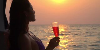 慢动作肖像美丽的亚洲女人在黑色比基尼和太阳镜享受与游艇在夏季日落饮酒。微笑性感女孩放松和享受奢侈的户外生活方式在暑假。