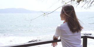 慢动作的年轻迷人的亚洲女人坐在阳台附近的岛屿海滩与风吹，看着美丽的大海。美丽的女孩在暑假放松和享受户外生活方式。