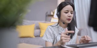 亚洲年轻女性戴着耳机在电话会议上讲话，用麦克风语音与客户互动，用键盘打字，在线客服与工作人员实时聊天，在家客厅办公