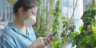迷人的亚洲女性休闲服戴口罩防毒手用智能手机数码设备在花园树在商场超市背景