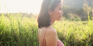 肖像亚洲美丽的年轻女子站在草地在绿色的自然与微笑的脸。放松女孩感到高兴，高兴和有快乐的时刻在假期在傍晚日落时间。