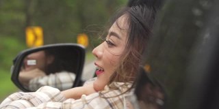 慢动作的快乐年轻美丽的亚洲女人旅行者坐在汽车与拉她的脸和手在雨天的车窗。漂亮女孩喜欢和享受户外生活方式和假期的乐趣。