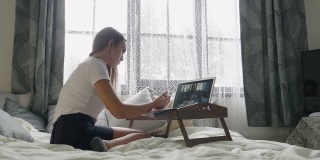 COVID-19大流行期间的在家教育。一名坐在卧室床上用笔记本电脑上在线课程的学生。少年学习。社会距离。