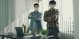 两名亚洲男子坐在他们的办公桌后面，展示着他们的自信，他们交叉着双臂，戴着面具看着镜头。