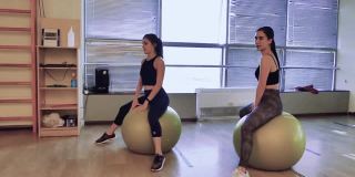 在健身房里，两个女孩正坐在健身球上，努力保持平衡。