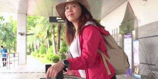 美丽的亚洲女人，一个长长的黑发，帽子，肩包和行李在手推车上等待机场候机楼寻找出租车，交通工具探索新的旅程。