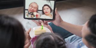 亚洲年迈的祖父母在社交距离中通过视频电话在家庆祝虚拟生日，感受被爱和快乐，由家人和侄子准备蛋糕。新标准的概念。