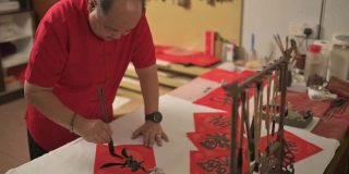 亚洲华人男性练习书法，为即将到来的中国新年庆祝家庭装饰目的与繁荣和良好的措辞写在一张红色的纸上