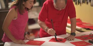 一位亚洲华人男子练习书法，为即将到来的中国新年庆祝家庭装饰目的繁荣和良好的措辞写在一张红色的纸上，并教他的女儿