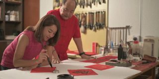 一位亚洲华人男子练习书法，为即将到来的中国新年庆祝家庭装饰目的繁荣和良好的措辞写在一张红色的纸上，并教他的女儿