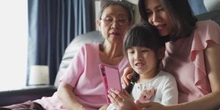 亚洲家庭的孩子、母亲、祖母在客厅里通过智能手机视频会议，开心地笑着聊天。快乐活动科技生活方式手机使用概念。