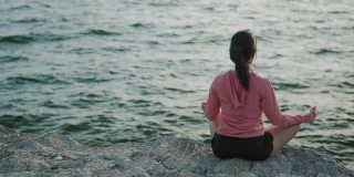 年轻美丽的运动女孩训练冥想坐在石头海边，而日落。