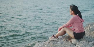 年轻女子跑步者坐在海边的石头上，从锻炼中休息，欣赏海滩日落的景色。