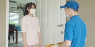新冠肺炎疫情期间，身穿蓝色制服、戴口罩和手套的亚洲快递员手持纸箱站在前门，以及一名接受快递员递送箱子的女性。