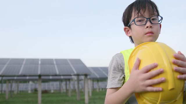 戴眼镜的男孩穿防护服和头盔。他双臂交叉站在太阳能电池板前。用于使用太阳能的可再生能源相关应用