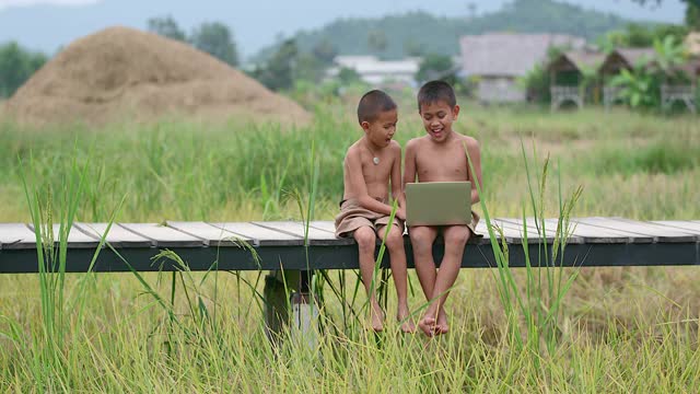 两个农村男孩愉快地玩着笔记本。在笑脸上在木桥上以田野为背景的接入方式，技术的到来在荒野中仍然可以使用互联网。