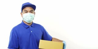年轻的亚洲快递员身穿蓝色制服，戴着防护面具和医用橡胶手套，拿着包裹纸板，竖起大拇指隔离。4 k决议。