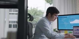 侧面的年轻迷人的亚洲泰国男子忙着工作的多屏幕电脑或智能平板电脑在桌子上在家里的自由职业者数据分析师，数据科学科学家的业务。