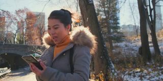 年轻的亚洲女性穿着冬衣微笑享受和平漫步在中央公园的冬季和黄色的树与地面的雪秋天手用智能手机设备的旅游概念