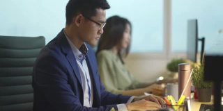亚洲华裔年轻商人在办公室里工作，在他的工作场所用笔记本电脑做他的报告
