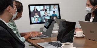 年轻的亚洲员工和资深的办公室员工团队通过电脑进行Zoom视频电话会议，商务人士共用办公桌使用电脑共同工作。