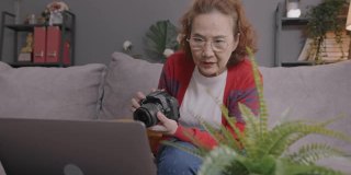 迷人的亚洲高级女子通过笔记本电脑学习在线摄影课程。坐在家里客厅的沙发上，用笔记本电脑和她的老师一起学习如何使用数码相机。