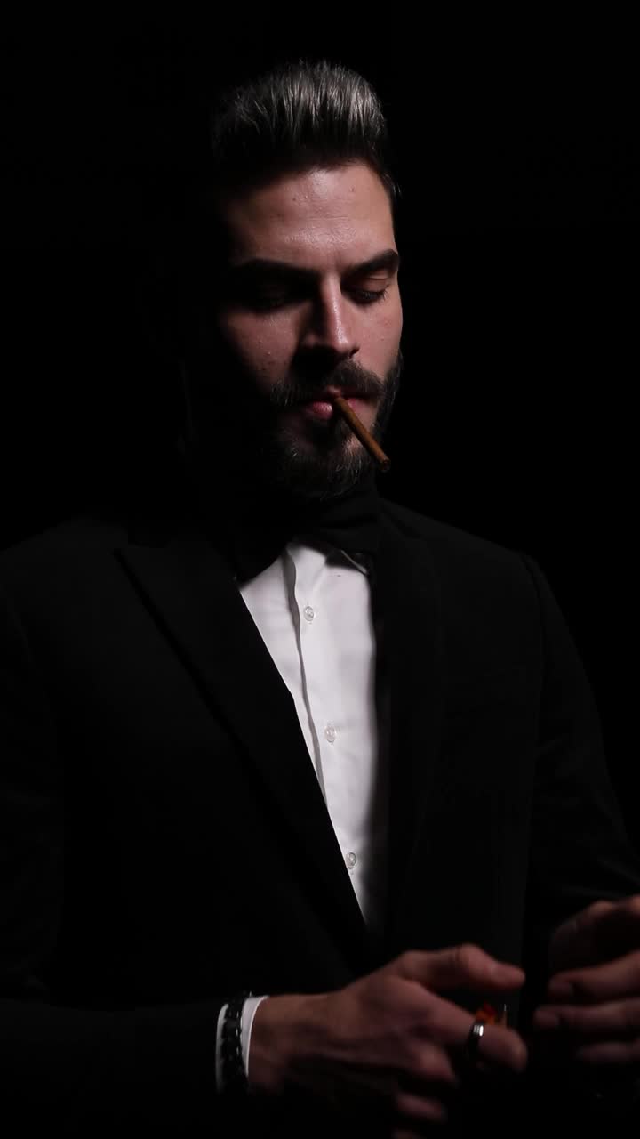 垂直肖像的戏剧性优雅的家伙点燃一根烟和吸烟，性感地看向一边和摆姿势，吸烟和离开黑暗在工作室的黑色背景