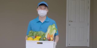 在居家隔离期间，一名亚洲快递员戴着口罩和手套，拿着装满食品、水果、蔬菜和饮料的杂货箱，站在客房前的货车旁。