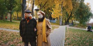老年配偶，戴着防护口罩的祖父母在秋季城市公园散步时交谈，冠状病毒大流行