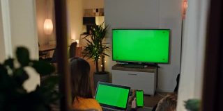 年轻夫妇使用笔记本电脑，智能手机和彩色键绿色屏幕的电视