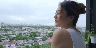 年轻美丽的亚洲女人在早上站在公寓阳台上，欣赏大都市的城市景观建筑的景色。2019冠状病毒病大流行封锁和心理健康概念。