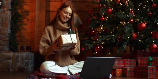 快乐的年轻女子拿着礼盒，而有视频电话上的笔记本电脑坐在圣诞树附近。