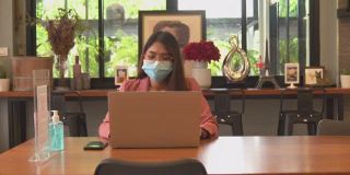 一位亚洲女性戴着外科口罩在咖啡店里用笔记本工作。