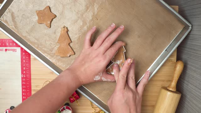 正上方，视角:亚洲中部妇女的手正在准备装饰的自制圣诞树饼干，姜饼，一颗星星在木制的圣诞桌子上通过挤压金属圣诞形状的模具在棕色面粉。