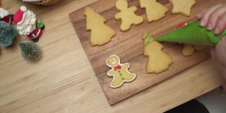 特写镜头:人的手用绿色糖霜来准备，装饰糖霜画体自制的圣诞树饼干、姜饼、圣诞树在木制圣诞桌子上。一些饼干在一个木盘子里