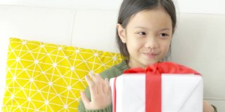 亚洲女孩在家里的沙发上拿着带红丝带的礼盒，假日概念。