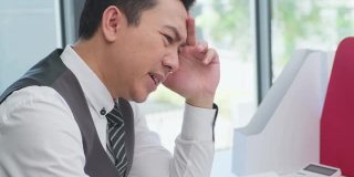 在办公室生病综合症，亚洲商人在电脑上工作有头痛，感到压力和生病的工作。商人对分配给他的工作感到灰心丧气。