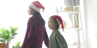 快乐的亚洲儿童戴着圣诞帽和毛衣一起在家里的窗户附近跳舞，圣诞概念。