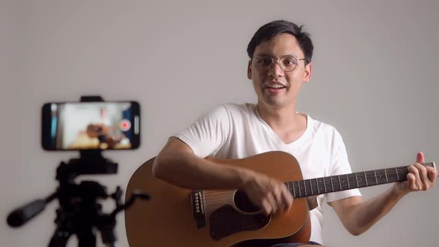一名亚洲男子弹吉他拍摄视频。男性用手机自拍录制吉他表演。专业吉他手在线铸造。