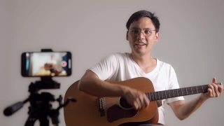 一名亚洲男子弹吉他拍摄视频。男性用手机自拍录制吉他表演。专业吉他手在线铸造。视频素材模板下载