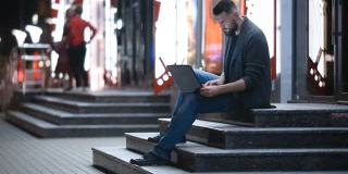 晚上，一个留着胡子的英俊男子坐在拥挤的街道上，把笔记本电脑放在腿上打字。自由工作的概念。聊天、发电子邮件、搜索或约会。