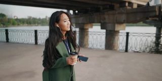微笑的亚洲女子走在河堤上，用智能手机听音乐，喝咖啡