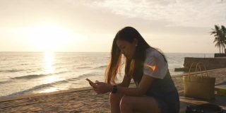亚洲女人使用智能手机，坐在海边的海滩在早上日出的天空背景。独自快乐旅行的概念。4 k慢动作。