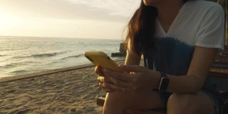 亚洲女人使用智能手机，坐在海边的海滩在早上日出的天空背景。独自快乐旅行的概念。4 k慢动作。