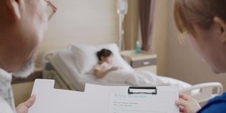 背景:亚洲家长和护理人员解释费用服务财务账单。可爱的小病人躺在病房的床上检查身体。儿童医院儿科病房。