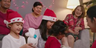 亚洲少女惊喜礼物盒给她可爱的哥哥和妹妹，而庆祝她的家人在圣诞节假期在家里的客厅。四个兄弟姐妹拥抱和微笑。
