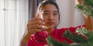 近距离亚洲少女享受装饰圣诞树在家里的客厅在圣诞节的假期。美女挂红球。