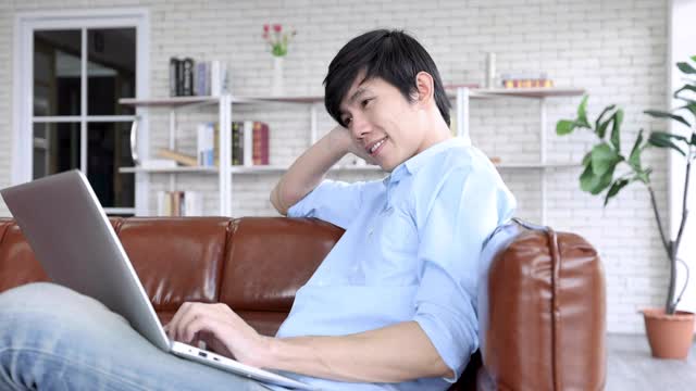 在家工作，微笑的亚洲人在客厅使用笔记本电脑。