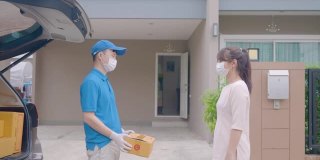 新冠肺炎疫情期间，身穿蓝色制服、戴口罩和手套的亚洲快递员手持纸箱站在前门，以及一名接受快递员递送箱子的女性。